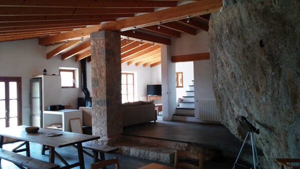 Un interior rústico con vigas de madera a la vista, un pilar de piedra, una pared de piedra, una chimenea, un televisor y una zona de comedor con bancos y una mesa.