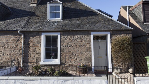 Een stenen gebouw met een klein raam en een deur waarop ‘Clava Cottage’ staat.