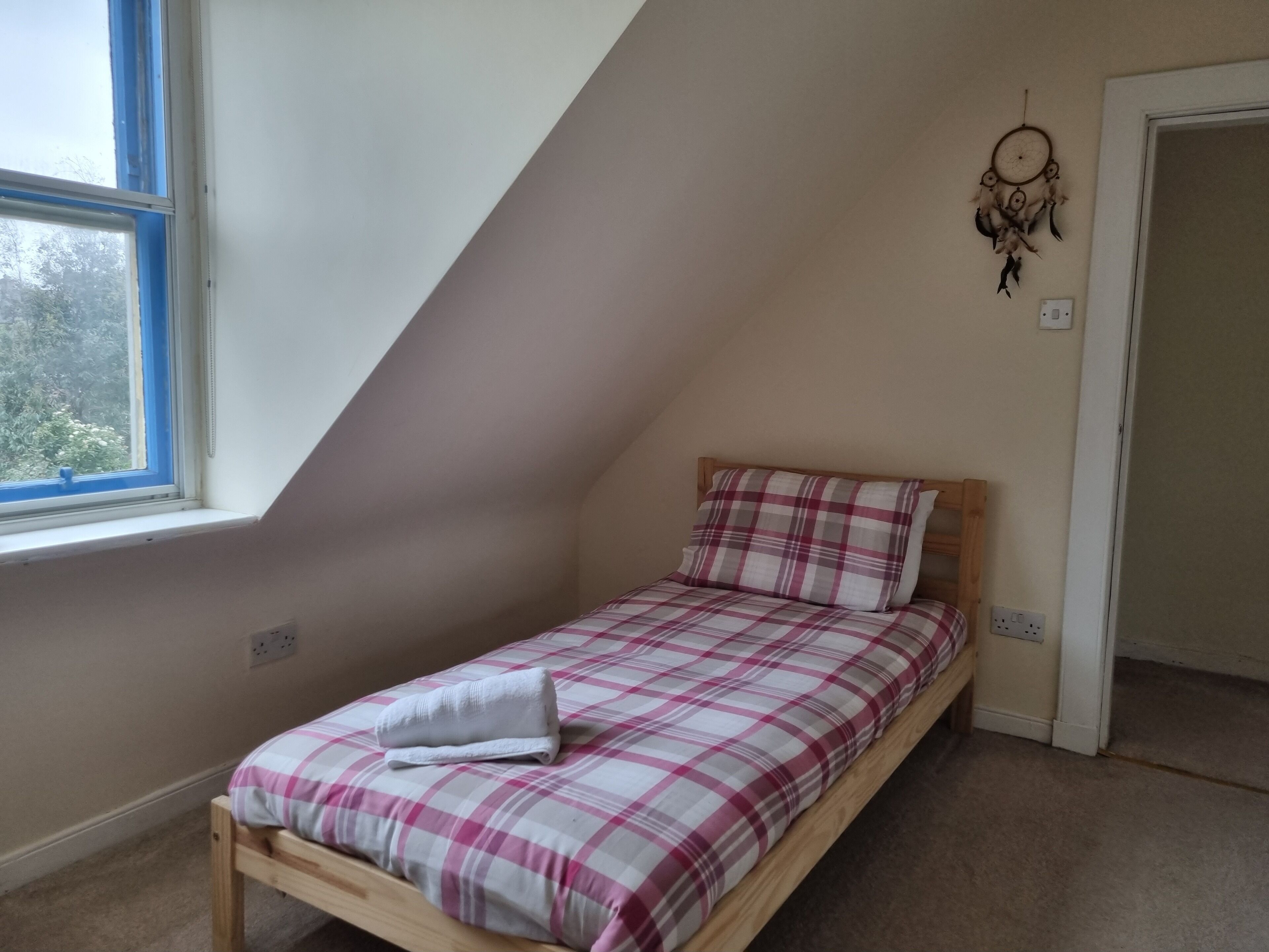 Een kleine kamer met een enkel bed, een schuine kap, een raam en een dromenvanger aan de muur.