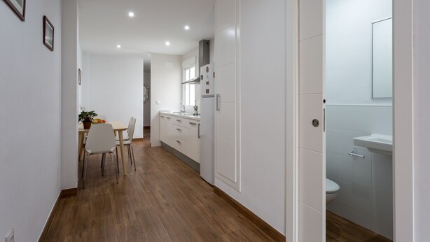 A modern kitchen with white cabinets, a dining area with a table and chairs, and a visible bathroom with a sink and toilet.