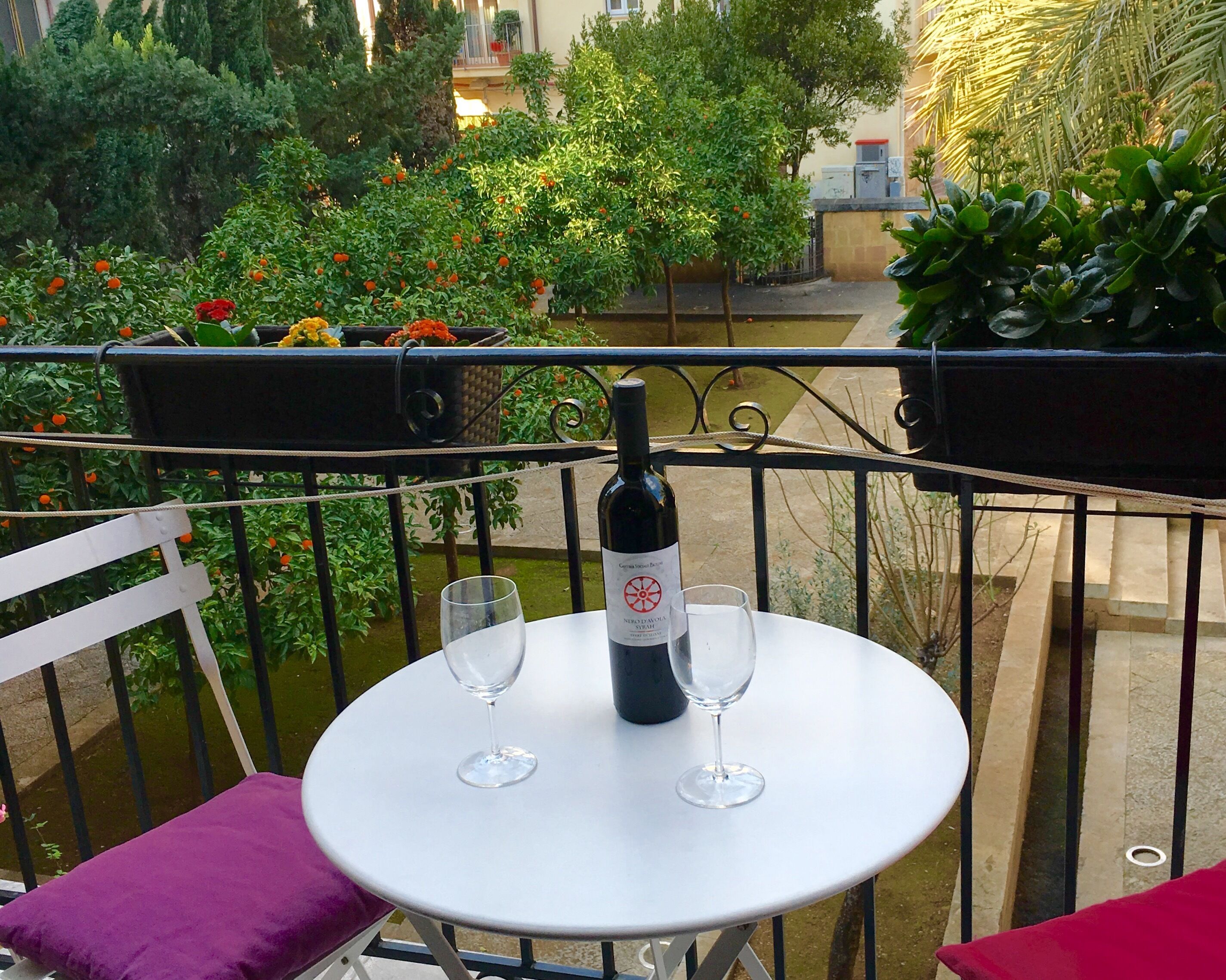 Une table ronde avec une bouteille de vin et deux verres, placée sur un balcon d’où l’on aperçoit un jardin et, au loin, un bâtiment.