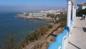 Vista costera con un edificio blanco de múltiples arcos, barandal azul y blanco y cielo azul despejado.