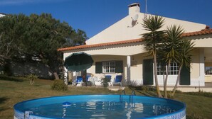 Una piscina con un borde decorativo, una casa con techo de tejas y un césped con sillas y sombrillas.