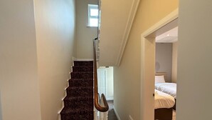 A staircase with a dark carpet, a white handrail, and a door leading to a bedroom with a bed and a window. - The Townlets Townhouse - 5BR - sleeps 13 (Belfast)