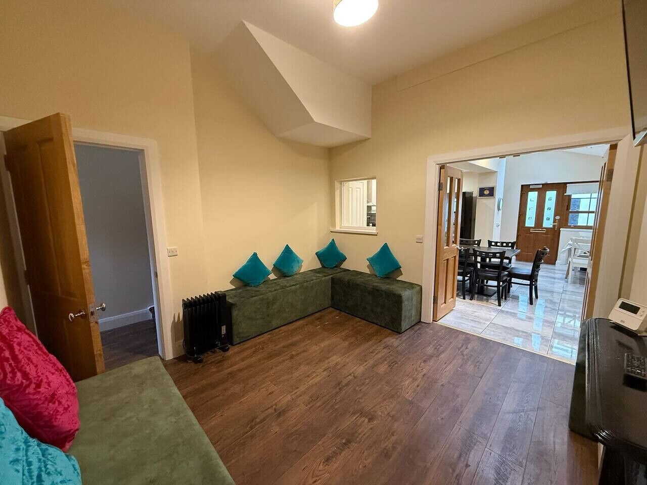 Un salon comprenant un canapé d’angle, un radiateur, un coin repas avec une table et des chaises, et un couloir menant à une autre pièce.