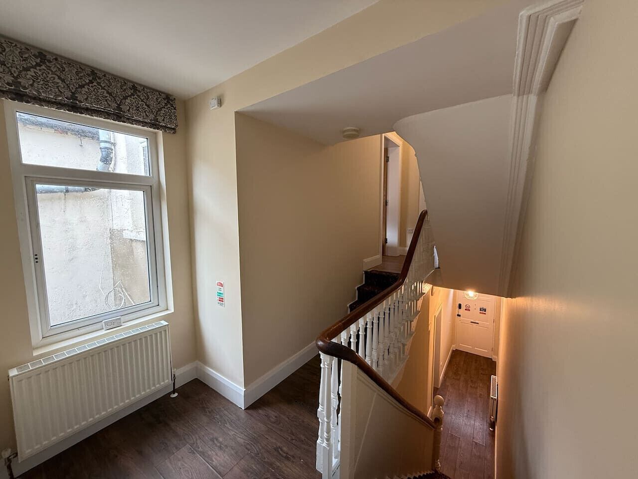 A staircase with a wooden handrail and white balusters, leading to a landing with a radiator and a window with a decorative valance.