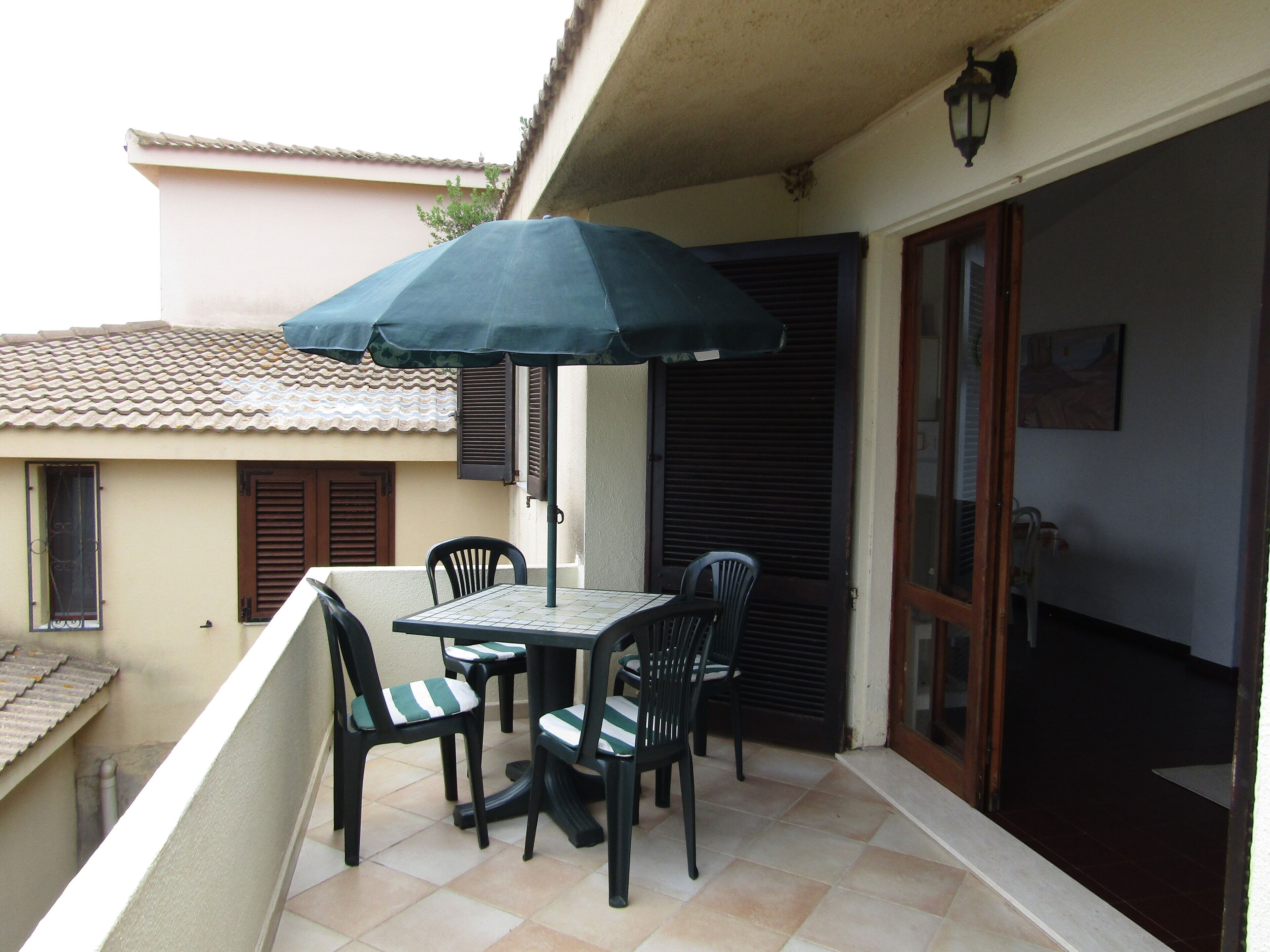 Un balcon avec une table et des chaises, un parasol vert, et une vue sur la rue et les bâtiments voisins.