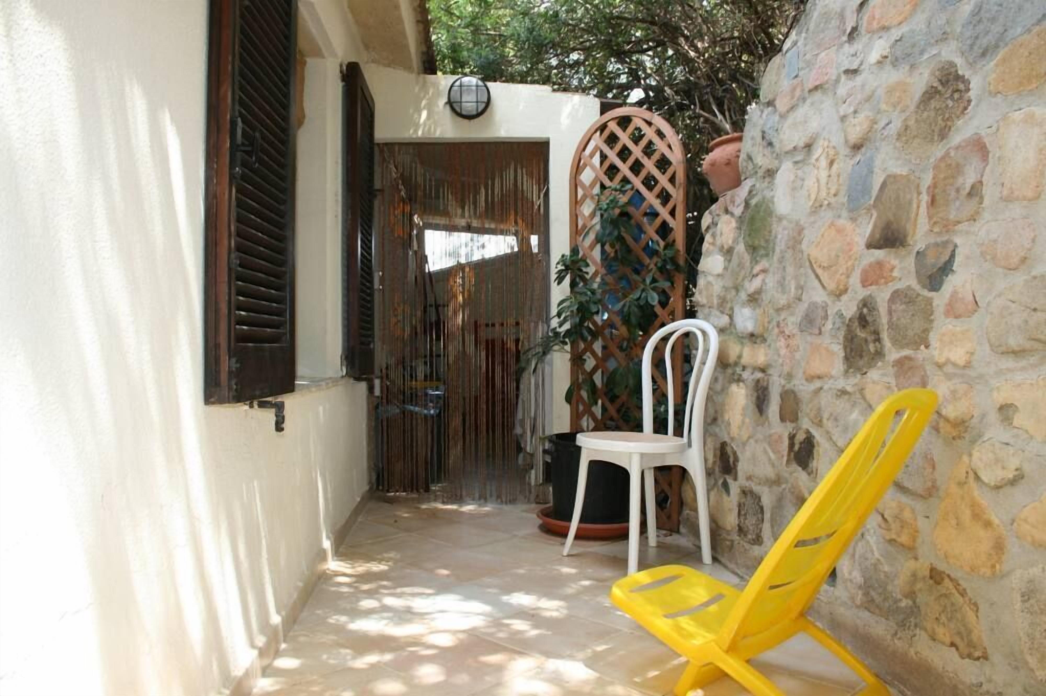 A narrow outdoor corridor with a white chair and a yellow chair, a stone wall, and a wooden lattice gate.