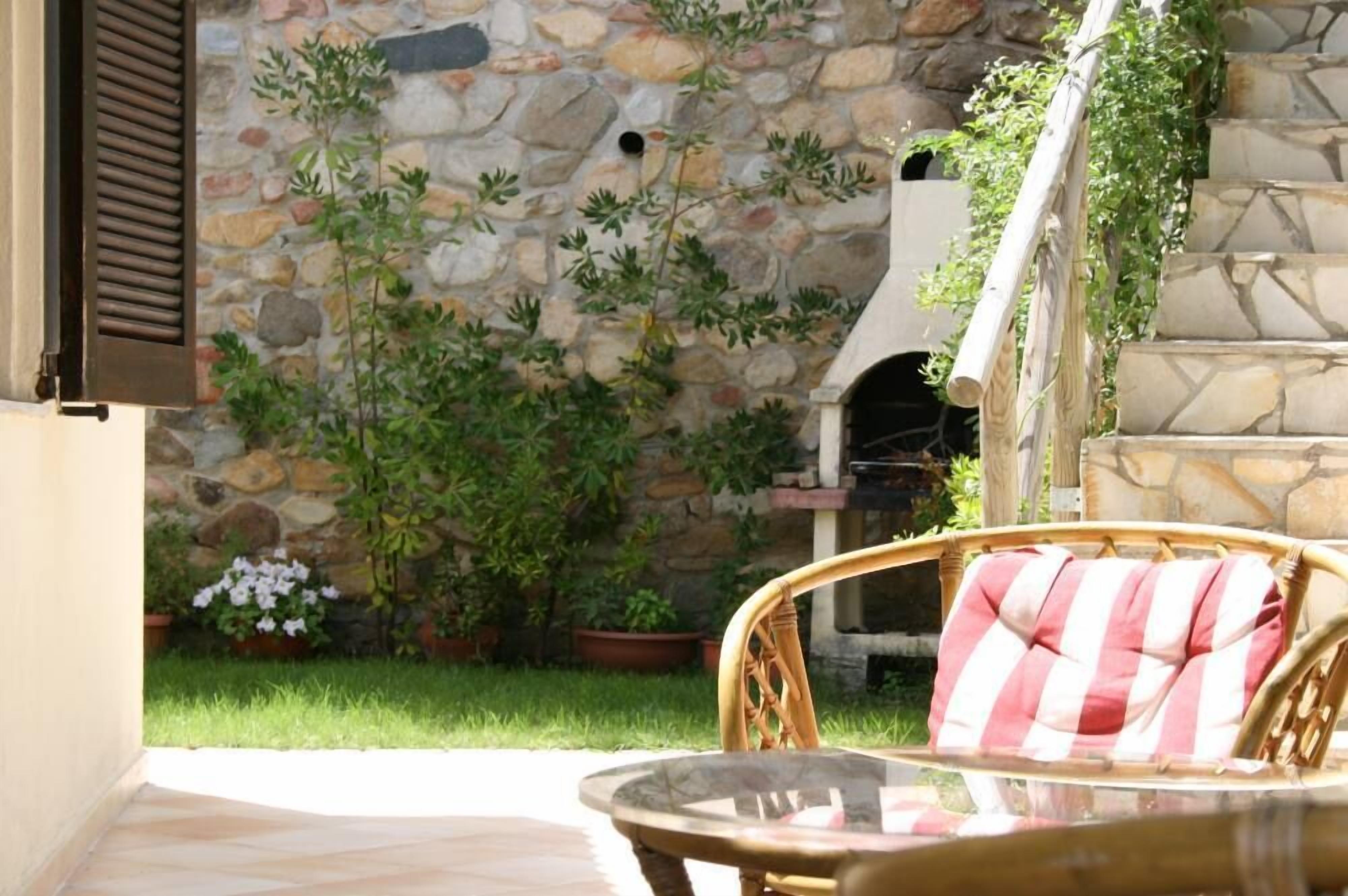 Un muro de piedra con plantas en macetas, un sillón de mimbre con cojín a rayas y una zona de barbacoa.