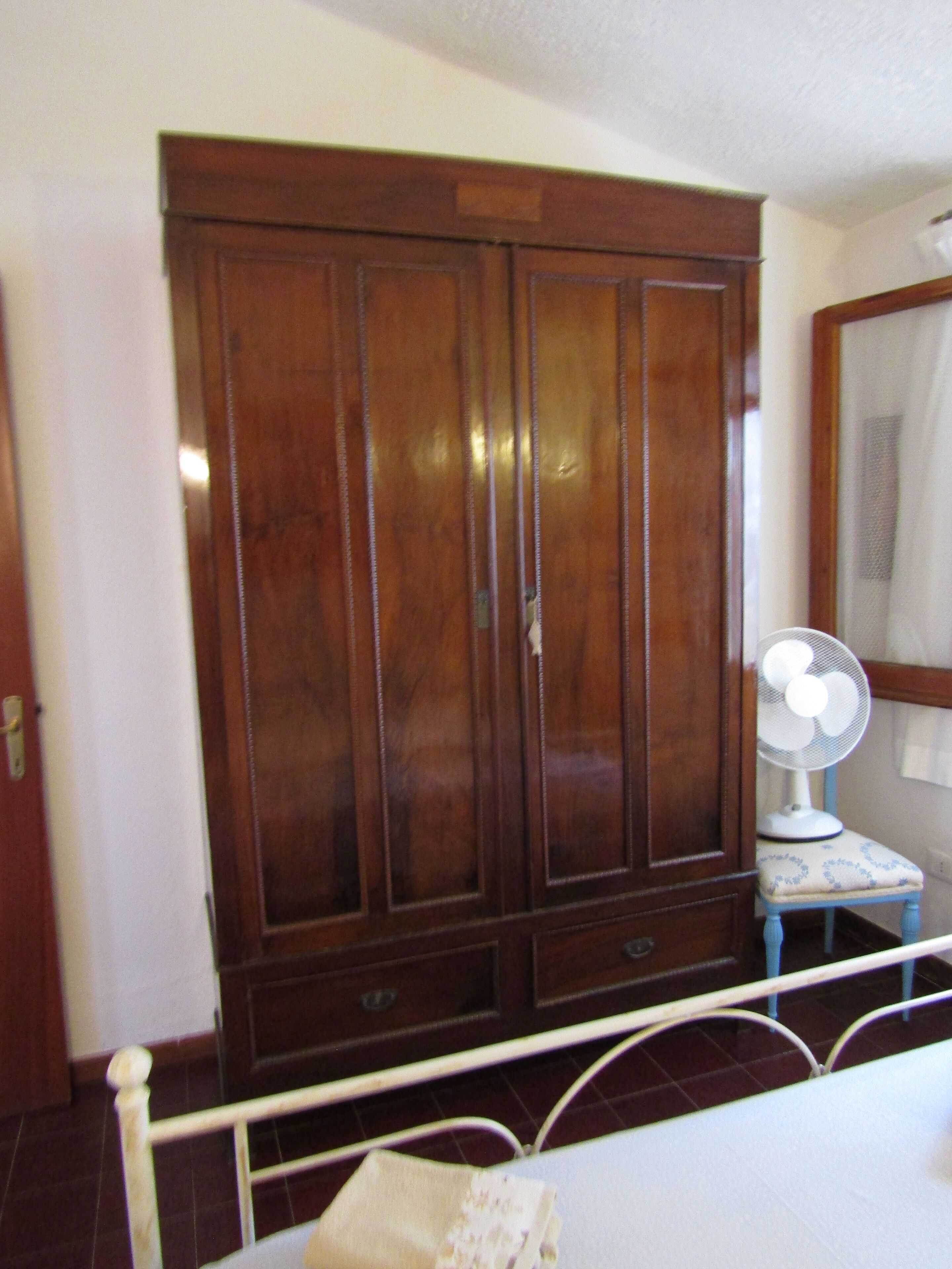 Une chambre avec une armoire en bois, une petite table de chevet avec un ventilateur et un lit doté d’une tête de lit blanche.
