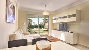 A modern living room with a sofa, a dining table with a bowl, a TV on a cabinet, and a view of a garden through a glass door.