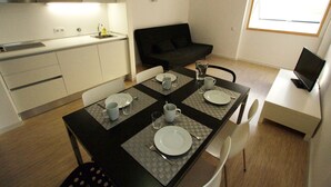 Eine kompakte Küche mit Mikrowelle, ein Esstisch für vier Personen mit Unterlagen und ein schwarzes Sofa im Wohnbereich.
