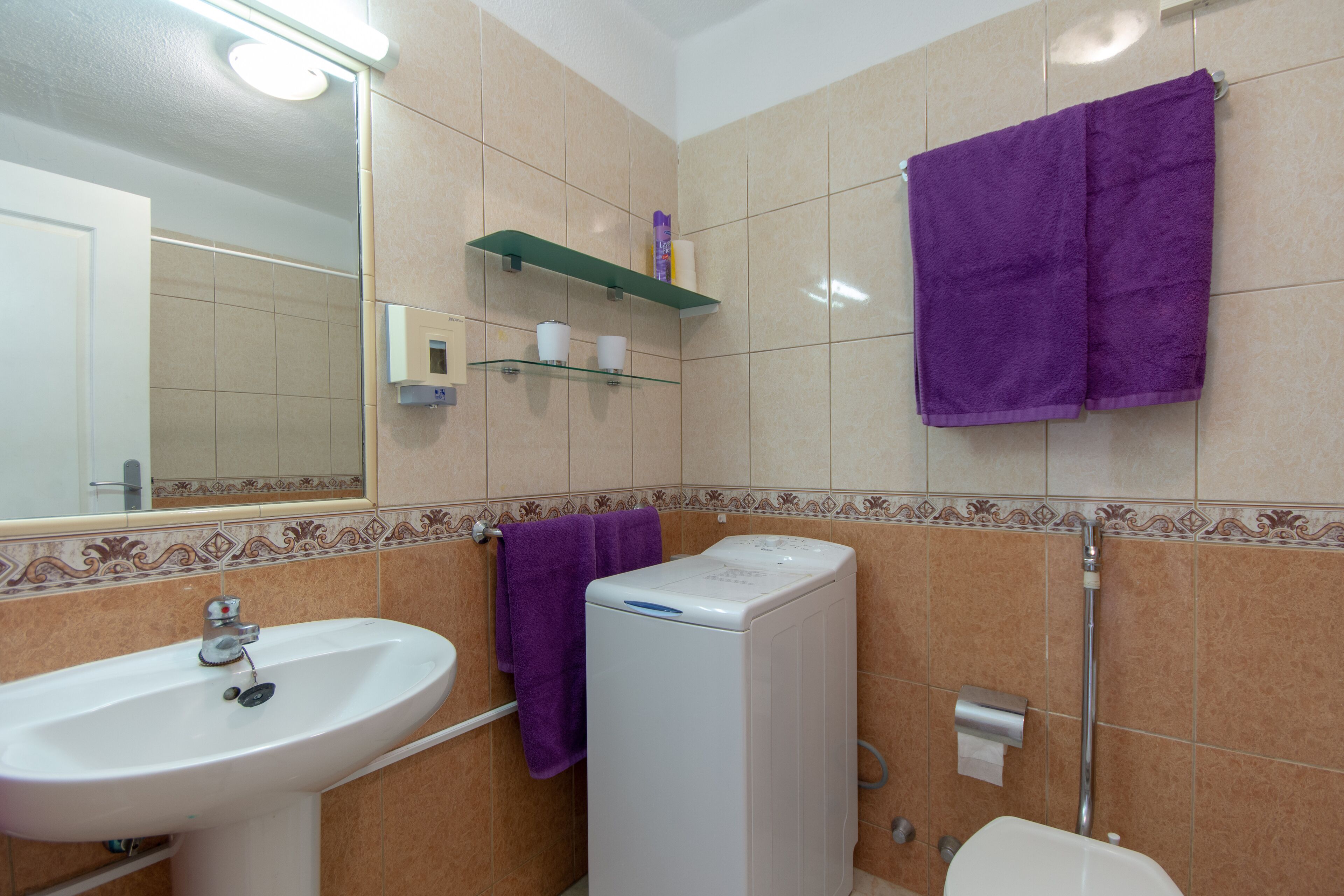 Un baño con lavamanos, lavadora y retrete. Hay toallas moradas colgadas en la pared y un estante con artículos de aseo.