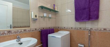 Un baño con lavamanos, lavadora y retrete. Hay toallas moradas colgadas en la pared y un estante con artículos de aseo.