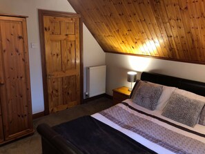 A bedroom with a sloped ceiling, a wooden wardrobe, a bedside table with a lamp, and a bed with multiple pillows.