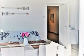 A modern dining area with a white table set for two, a vase with pink flowers, and a wall-mounted light fixture.