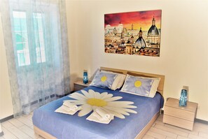 A hotel room with a bed featuring a large floral bedspread, a cityscape painting on the wall, and a window with sheer curtains.