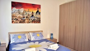 A hotel room with a bed featuring a blue bedspread with a large white daisy design, white pillows, and neatly folded towels. A painting of a cityscape is on the wall above the bed.