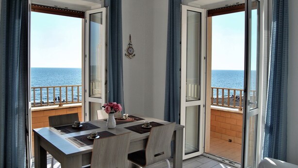 A dining area with a table and chairs, a vase with flowers, and a view of the sea through open doors.