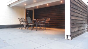 A covered outdoor dining area with a table and chairs, a wooden wall, and a white building in the background.