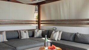 A sectional sofa with striped pillows, a wooden coffee table with a bottle and watermelon, and a view of a building through a window. - CELESTINE, beach front villa with roof top pool and spectacular views. (Kolymvari)