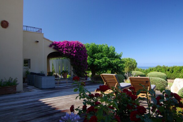 A wooden deck with two wooden lounge chairs, a potted plant with red flowers, and a view of the sea.