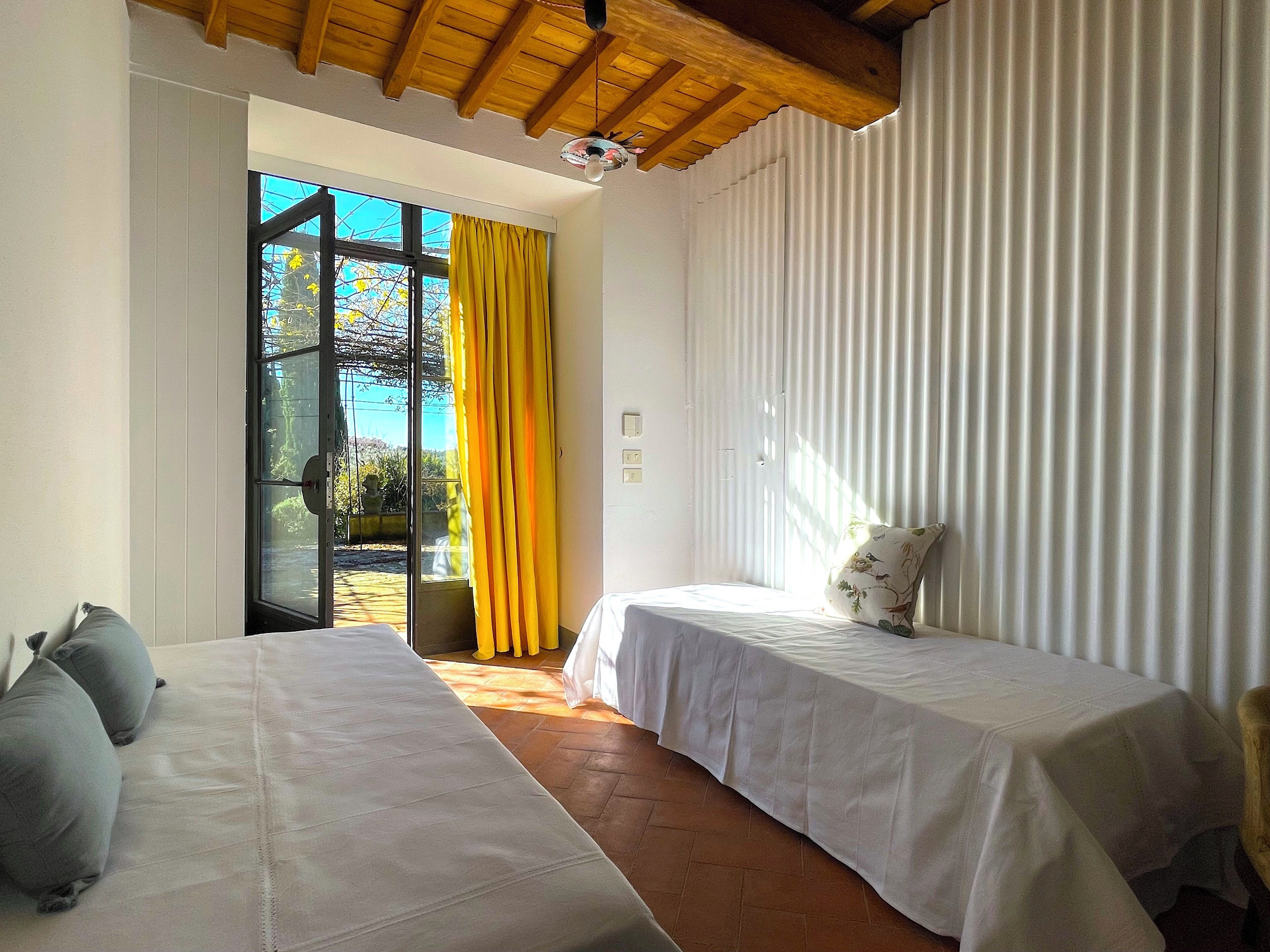 Habitació amb dos llits, sostre de fusta, finestra gran amb cortines grogues i vista a vegetació a l'exterior.