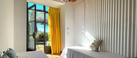 Habitació amb dos llits, sostre de fusta, finestra gran amb cortines grogues i vista a vegetació a l'exterior.