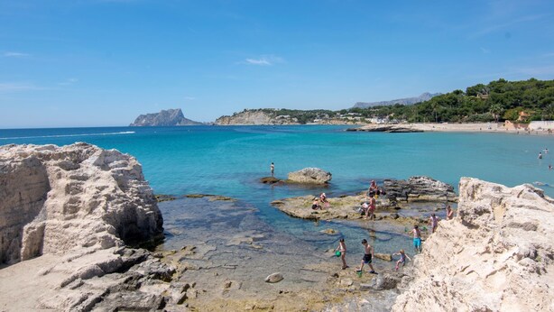 A rocky beach with clear turquoise water, people enjoying the beach, and a mountainous backdrop.