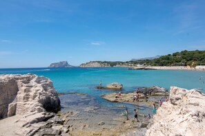 A rocky beach with clear turquoise water, people enjoying the beach, and a mountainous backdrop.