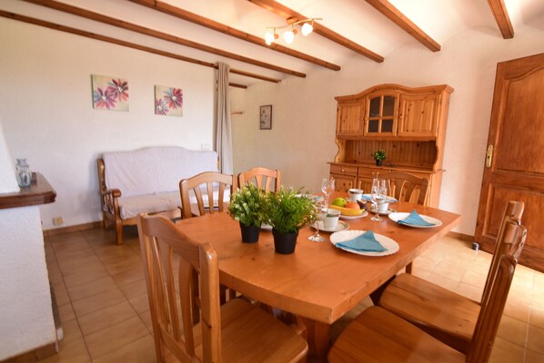 A dining area with a wooden table set for four, surrounded by wooden chairs. There are potted plants on the table, and a wooden cabinet is visible in the background.