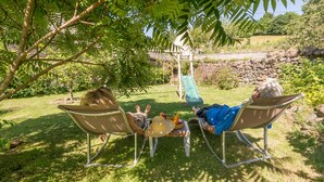 Dos personas descansando en sillas de jardín bajo un árbol, con una pequeña mesa entre ellas que sostiene bebidas y un sombrero.