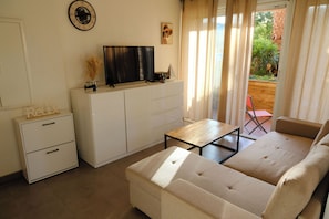 Uma sala de estar com sofá, televisão sobre um móvel, uma mesinha e vista para um jardim através de uma janela.