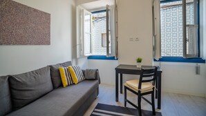 Ein kompakter Wohnraum mit grauem Sofa, einem kleinen Tisch und zwei Fenstern mit blauen Fensterrahmen.