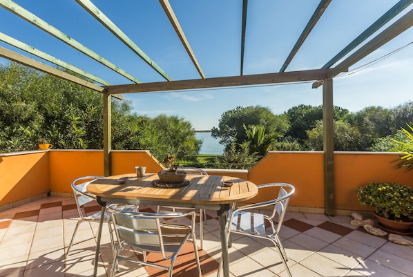 An outdoor patio with a wooden table and chairs, surrounded by orange walls and a view of trees and water. - home holiday sea view in Mandriola (San Vero Milis)