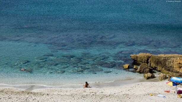 Una playa con agua cristalina, un promontorio rocoso y una persona sentada en la arena.