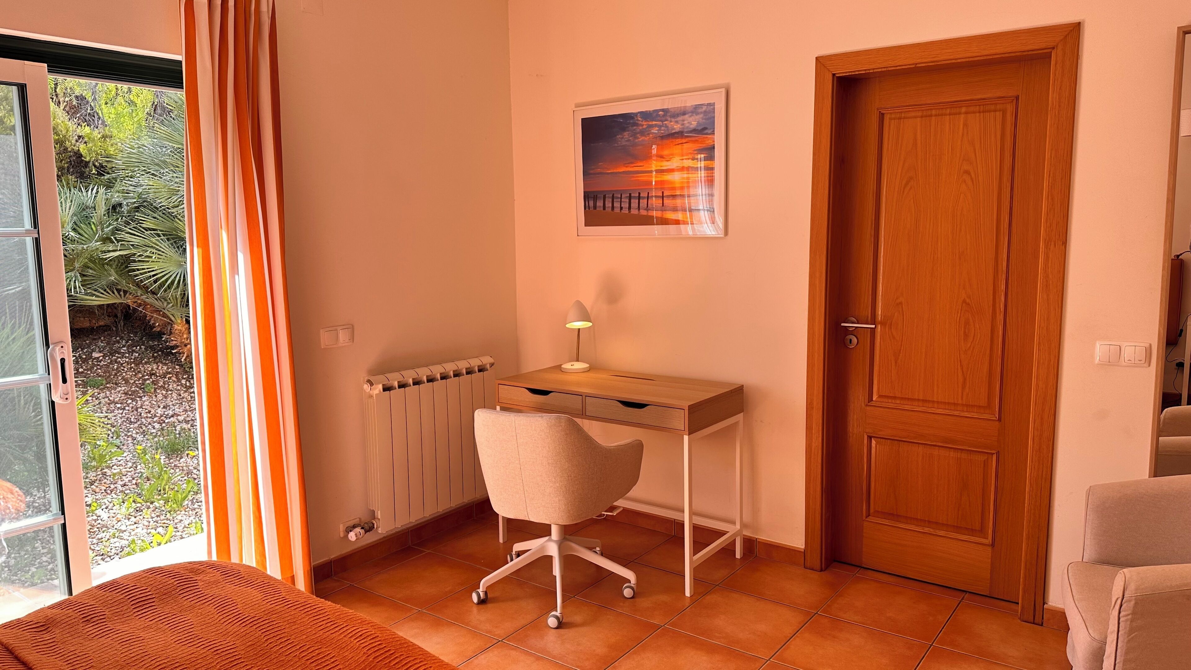 A room with a wooden desk, a chair, a radiator, a door, and a window with curtains.