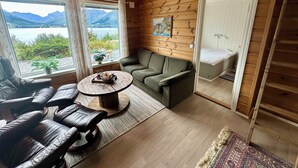 Una cabaña acogedora con interior de madera, un sofá, una mesita de centro y vistas a montañas y un lago a través de amplios ventanales.
