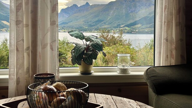 Una habitación con vista a montañas y un lago, una planta en maceta, un bol de pan y una vela en el alféizar de una ventana.