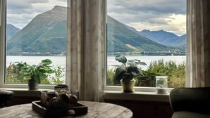 Habitación con un amplio ventanal con vistas a un lago y montañas. Hay plantas en macetas en el alféizar y una mesa de madera con una bandeja de galletas.