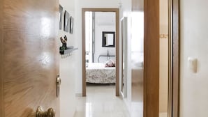 Un couloir mène à une chambre qui contient un lit, une commode et un miroir. Des étagères murales sont fixées au mur et accueillent des objets décoratifs.