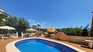 En swimmingpool med liggestole og parasoller, omgivet af fliseterrasse og grøn bevoksning.