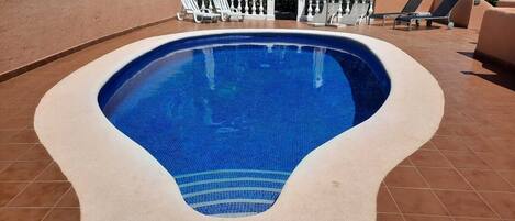 Una piscina de forma singular, rodeada por una solana con baldosas y barandillas blancas.