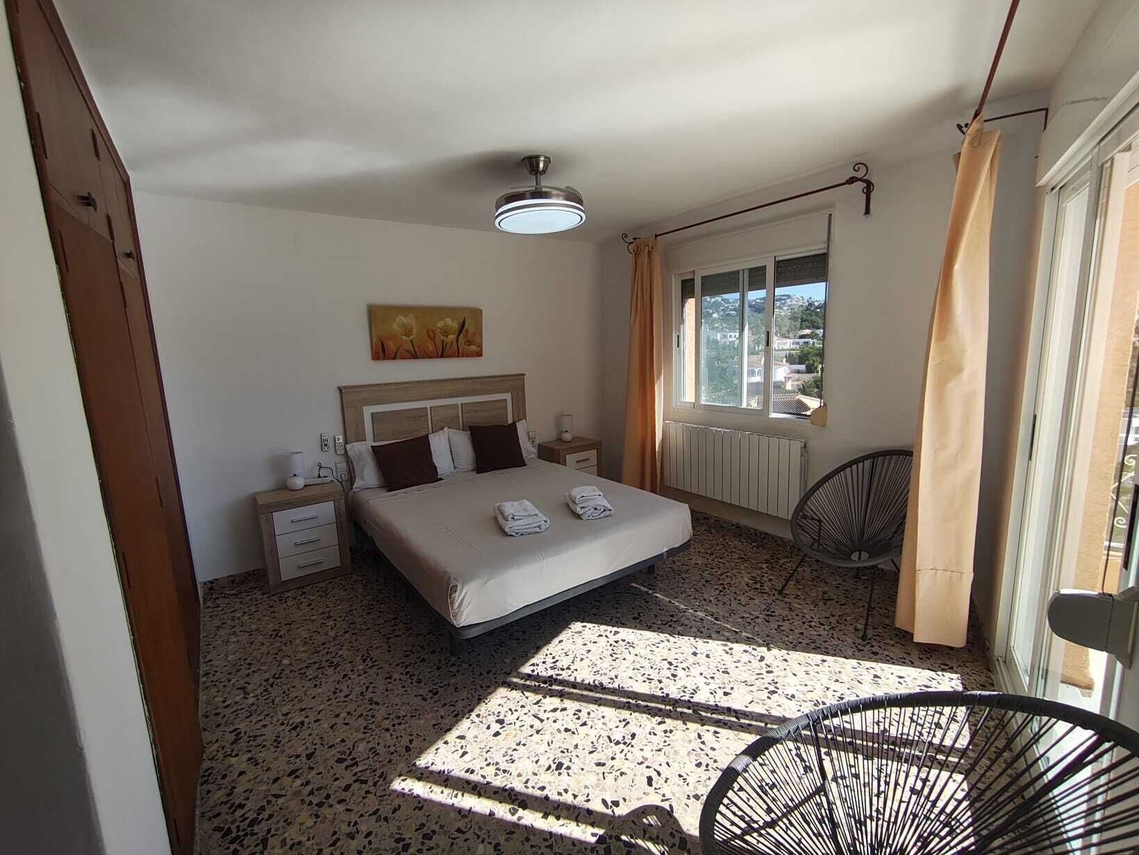 A bedroom with a bed, bedside tables, a ceiling fan, a window with curtains, and a radiator.