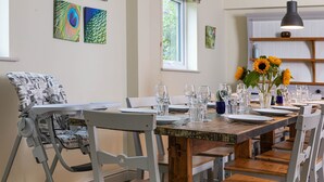 A dining area with a wooden table set for six, surrounded by white chairs, and a high chair with a patterned cushion. - Sleeps 8, 4 Bedrooms, Dogs Welcome, No Smoking, Wood burner, Wi-Fi*, Garden, Games Room (Cheltenham)