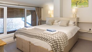 A bedroom with a large bed, a window with curtains, a bedside table, and a wall-mounted lamp. - Sleeps 8, 4 Bedrooms, Dogs Welcome, No Smoking, Wood burner, Wi-Fi*, Garden, Games Room (Cheltenham)