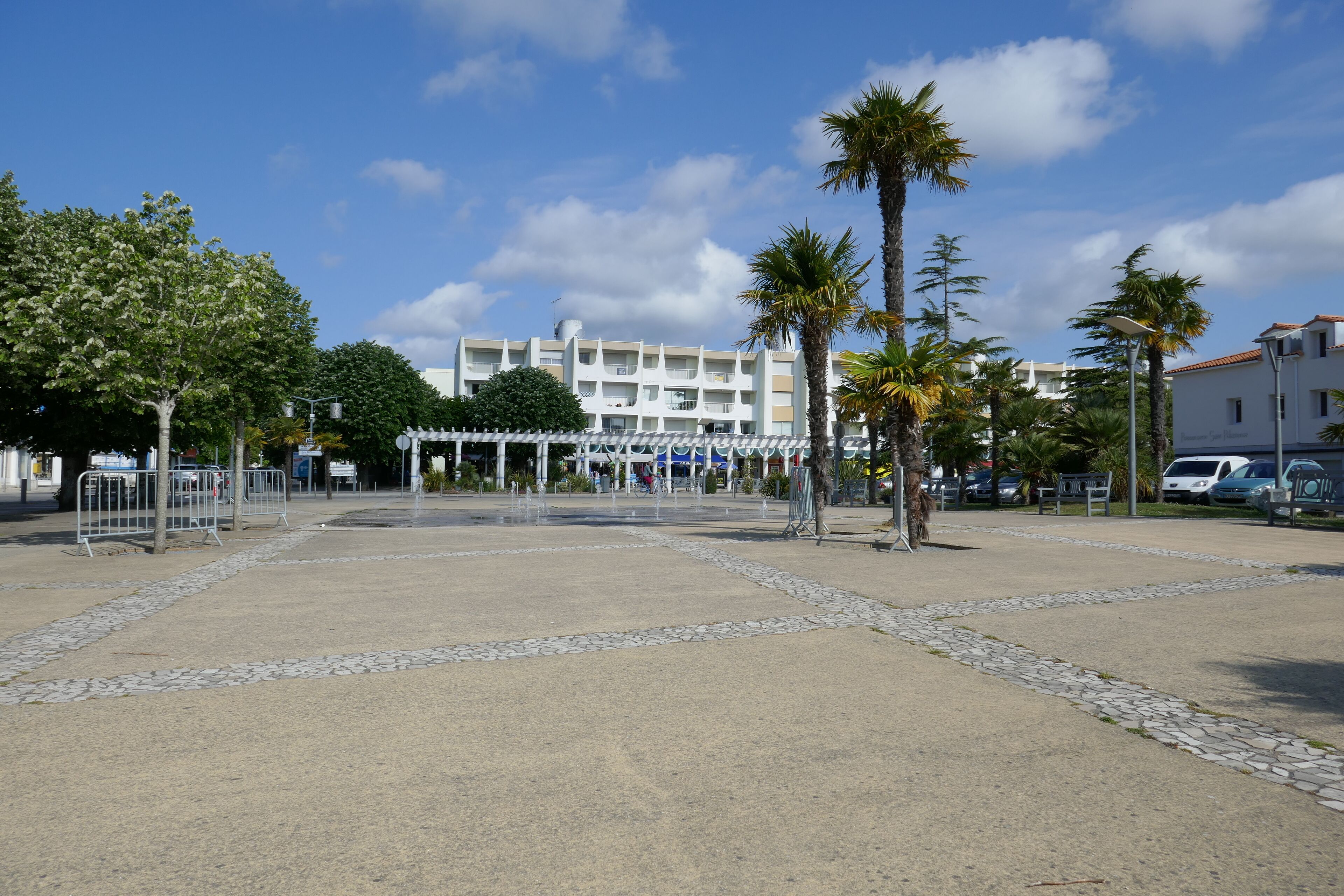 A spacious outdoor area with a paved walkway, palm trees, and a building with multiple balconies in the background.