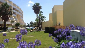 Une cour avec un terrain de jeux, des palmiers et des fleurs violettes.
