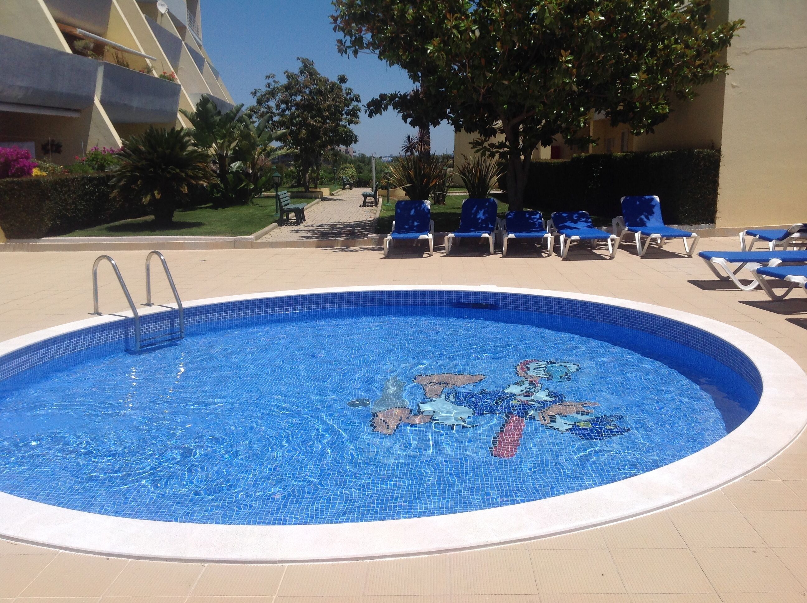 A swimming pool with a mosaic design, surrounded by lounge chairs and a paved deck.