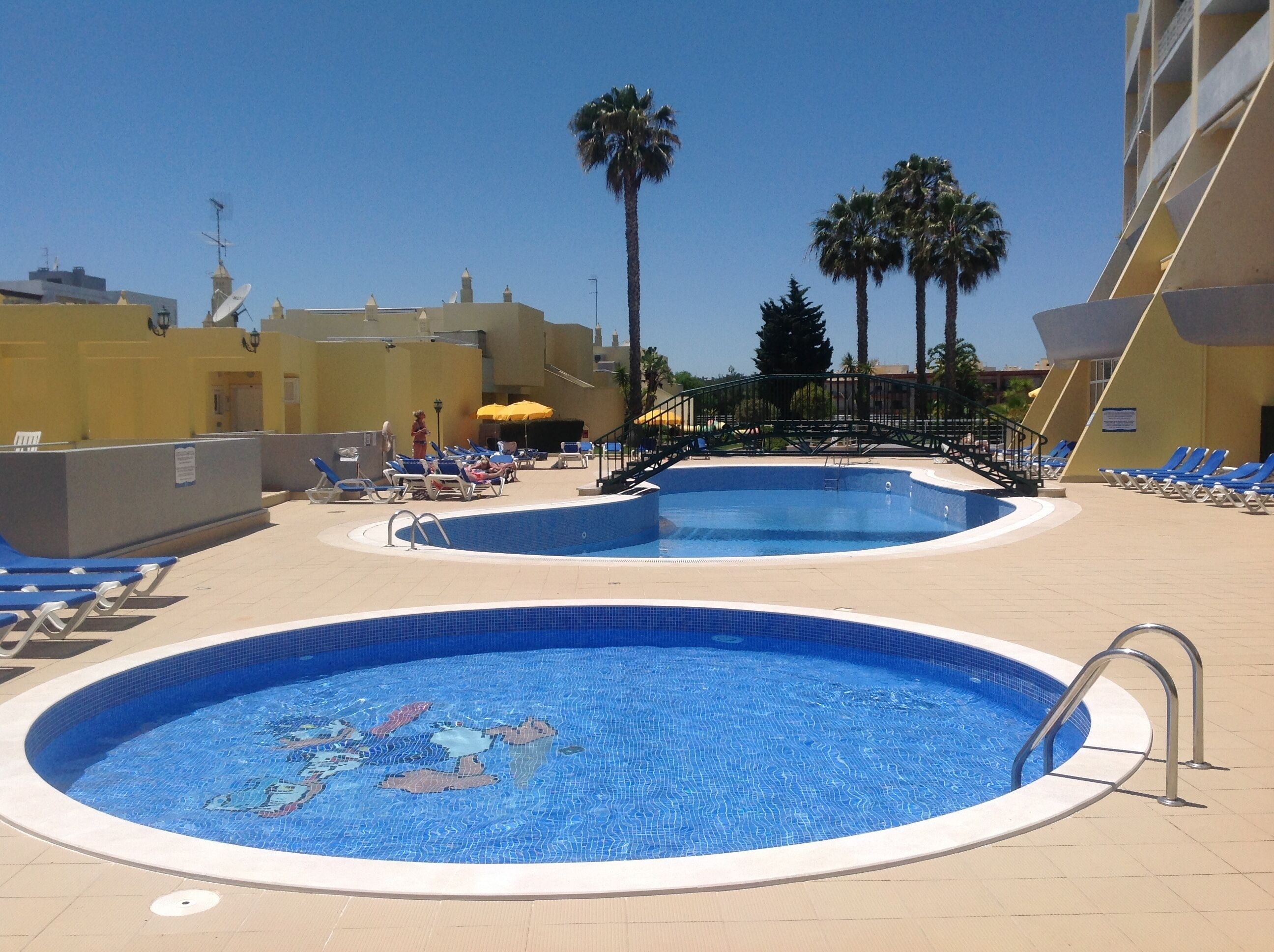 Un espace piscine comprenant deux piscines, des chaises longues et des palmiers.
